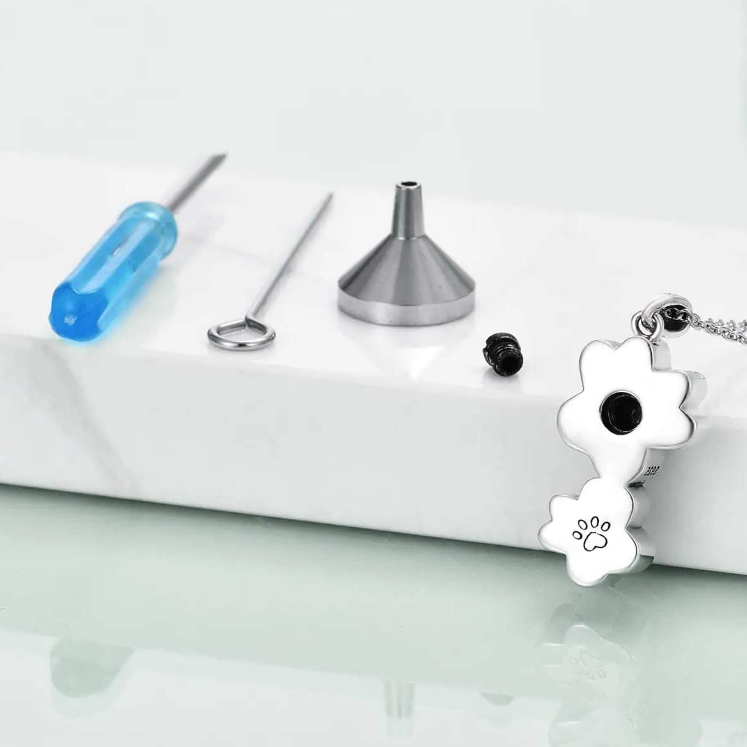 Parte trasera de colgante de plata para cenizas, donde se puede ver el orificio, el embudo, tuerca y destornillador para introducirlas. Con una huella de PANDORA grabada