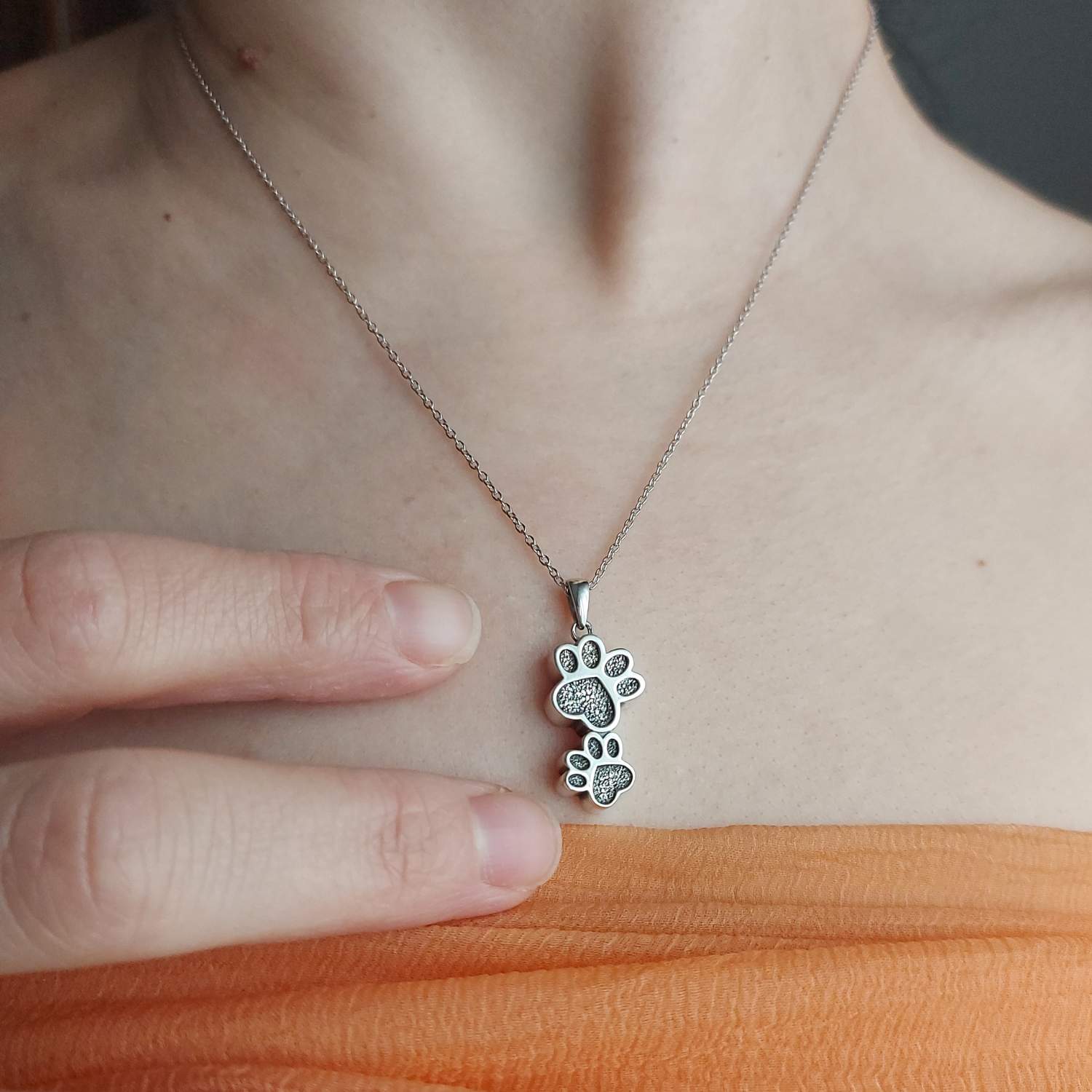 Collar con dos huellas de perro o gato en cuello de mujer, entre 2 dedos para que se vea el tamaño pequeño. Con camisa naranja del corte ingles