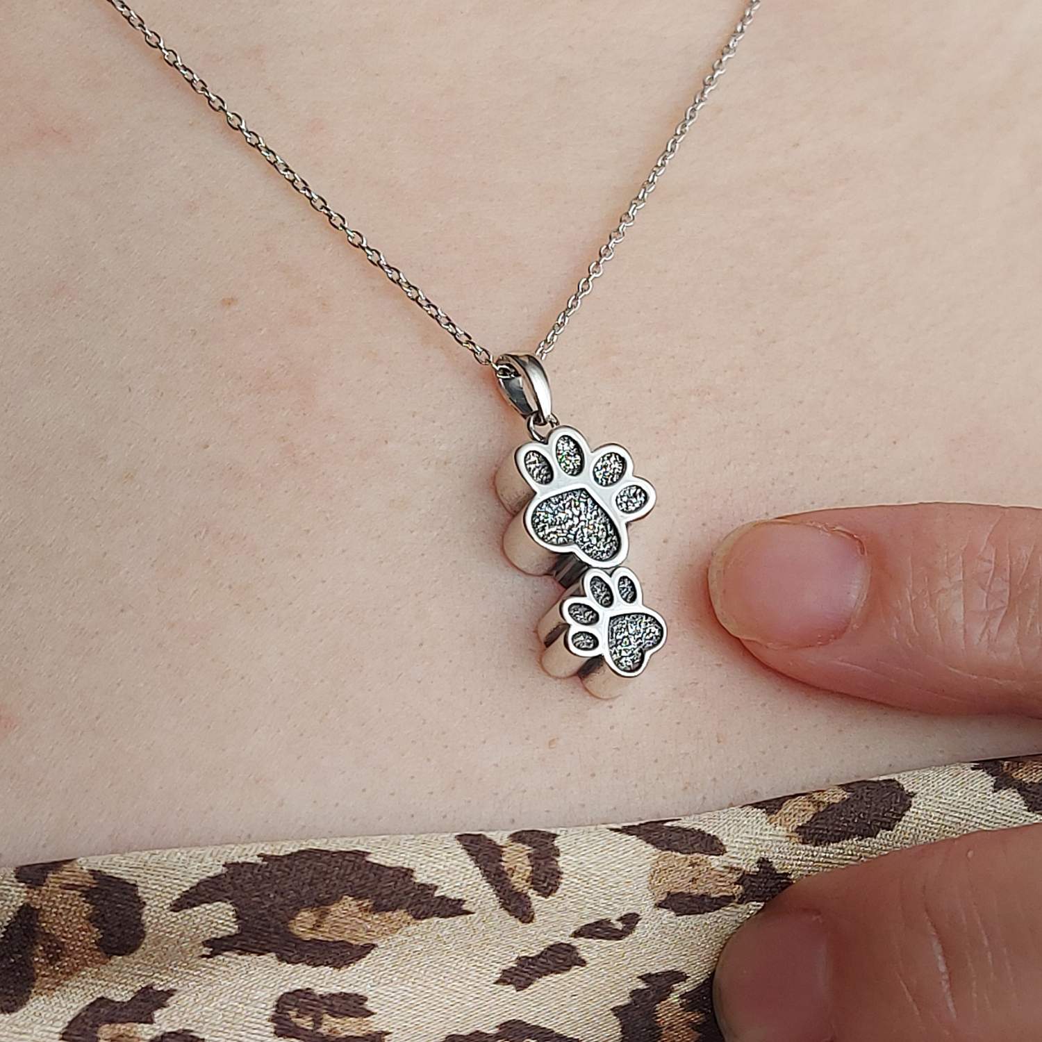 Foto de cerca del collar de 2 huellas de plata con capacidad para cenizas de mascota, sobre escote de modelo con camisa estampada del corte inglés. Señalando el colgante para que se aprecie su tamaño pequeño.