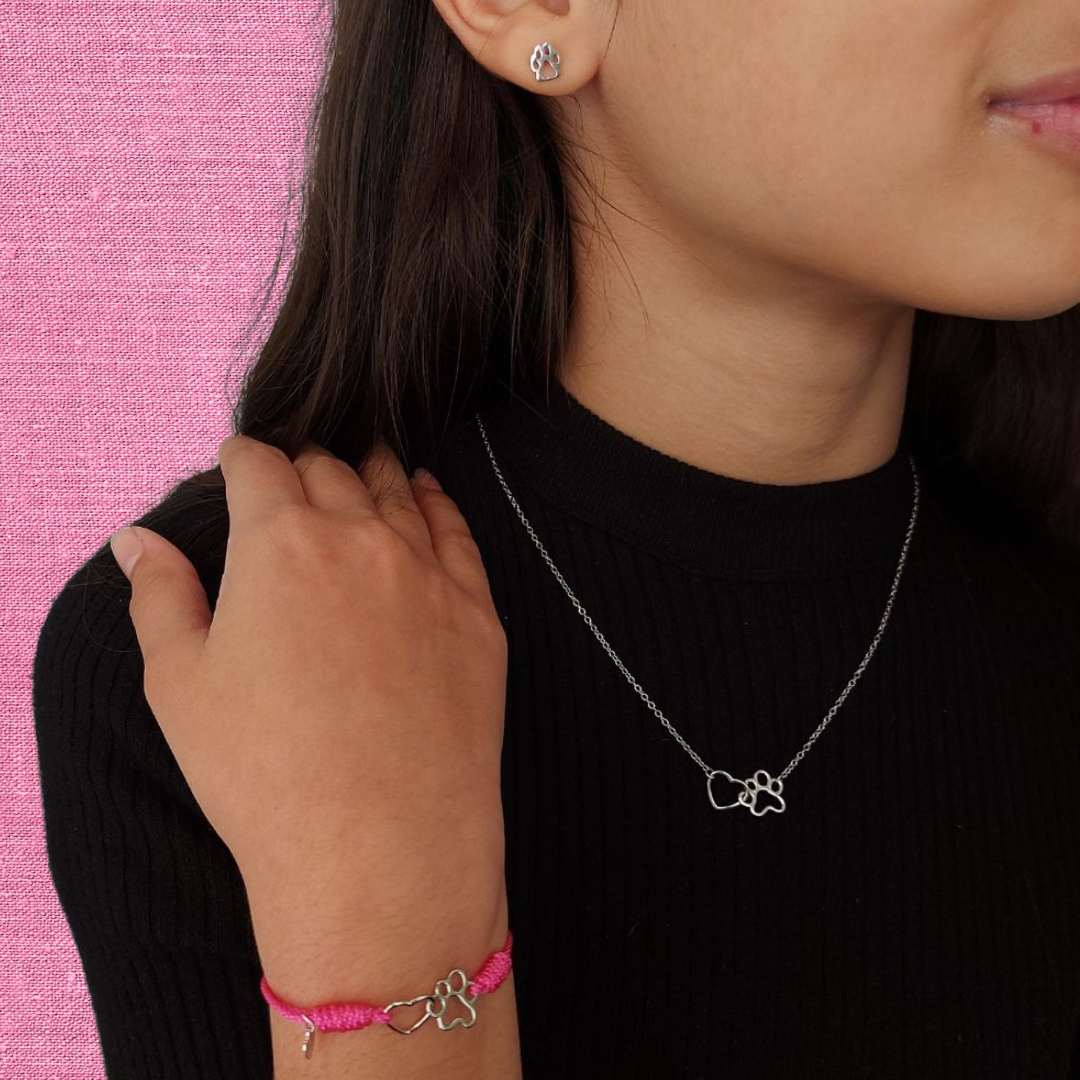 Niña con una pulsera de hilo rosa y ajustable con diseño de pata y corazón de perro o gato, collar de plata con el mismo colgante y unos pequeños pendientes de plata con huellita de mascota, conjunto de comunión de pandora