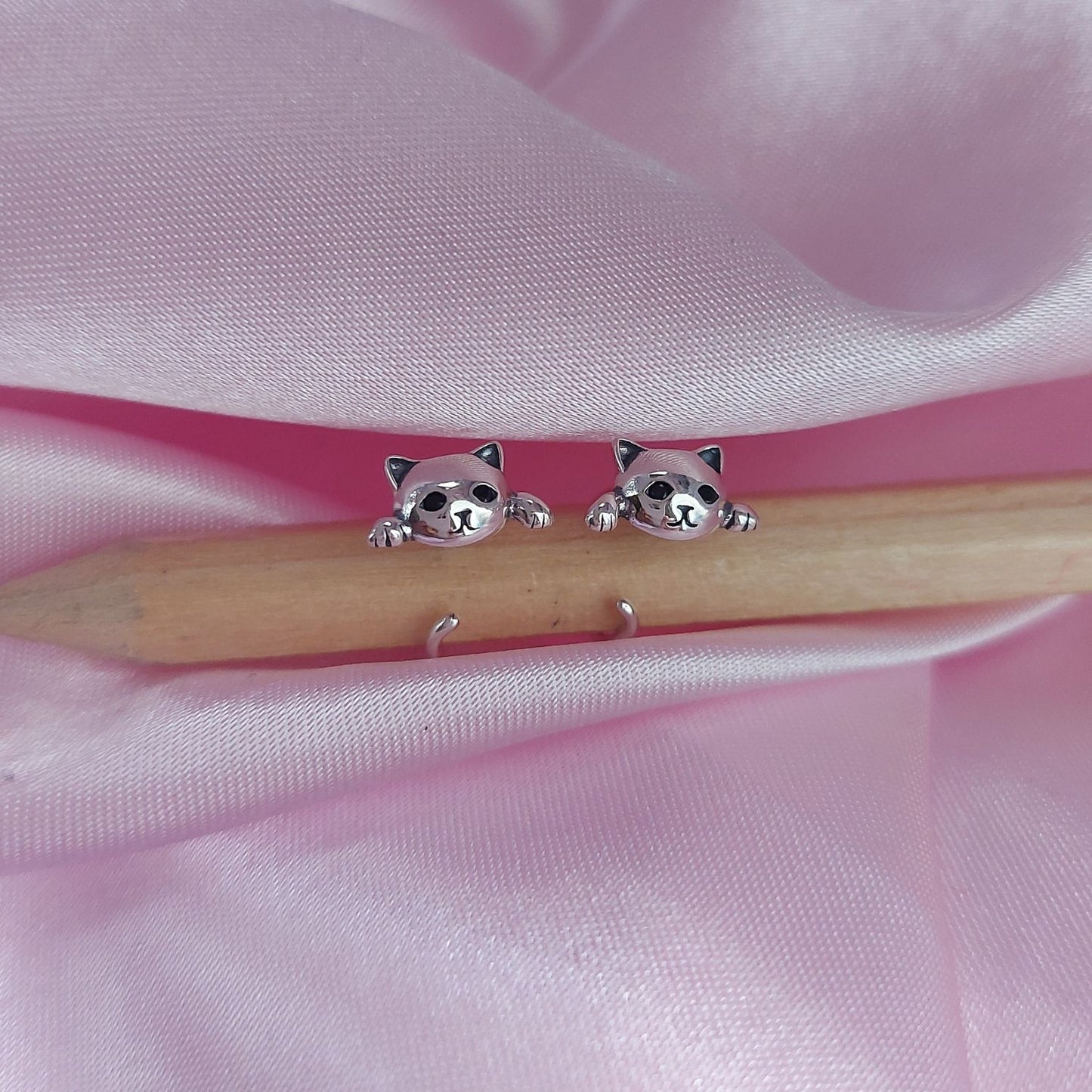 Pendientes con gatos expuestos agarrando a un lápiz de madera sobre tela rosa de sansaru