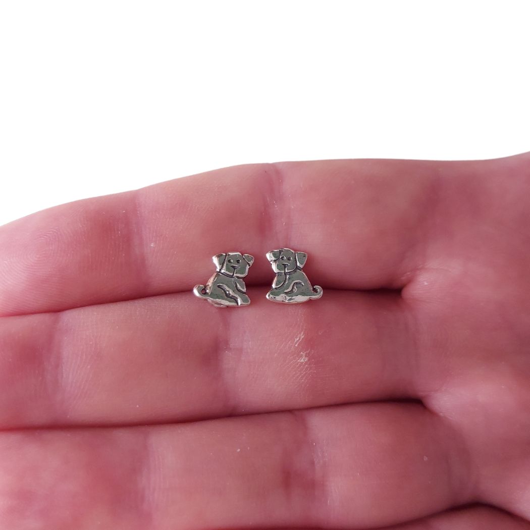Pendientes pequeñitos de perritos en plata de ley 925, colocados entre los dedos de una mano.