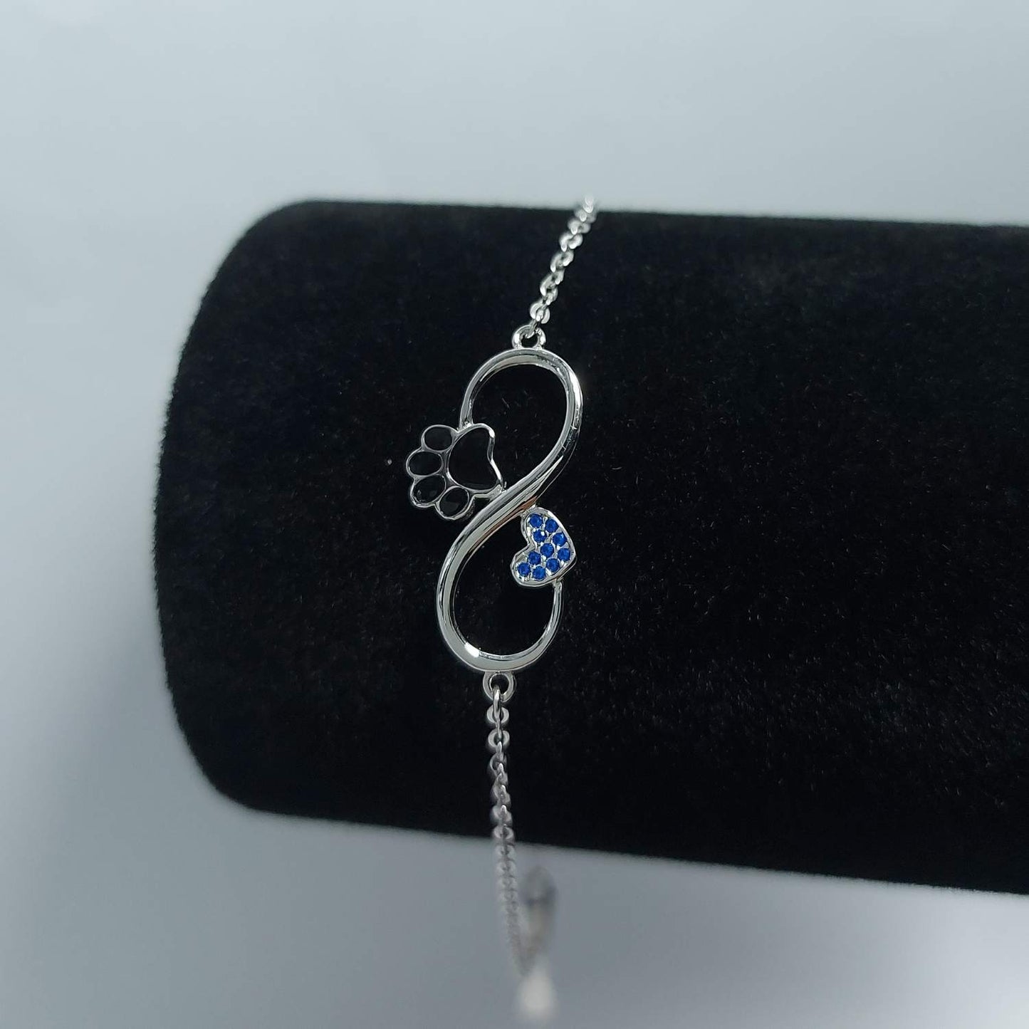 Pulsera con el símbolo del infinito de plata, con huella negra de mascota y corazón con brillantes azules incrustados, sobre expositor de pulseras negro de Pandora