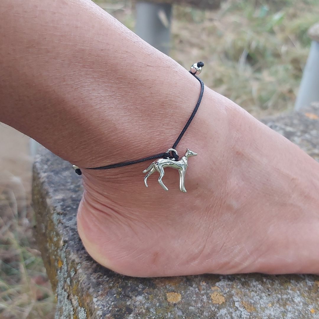 Tobillera de hilo negro con un colgante de un perrito galgo de plata de PANDORA, los nudos ajustables están protegidos por unas bolitas de plata y el pie está sobre un banco de piedra.