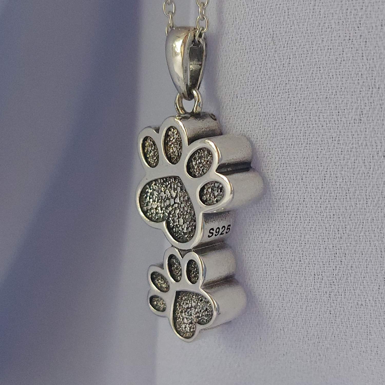 Foto al detalle del colgante para cenizas, donde se aprecia el relieve de las huellas con tonos negros y el sello de plata S925