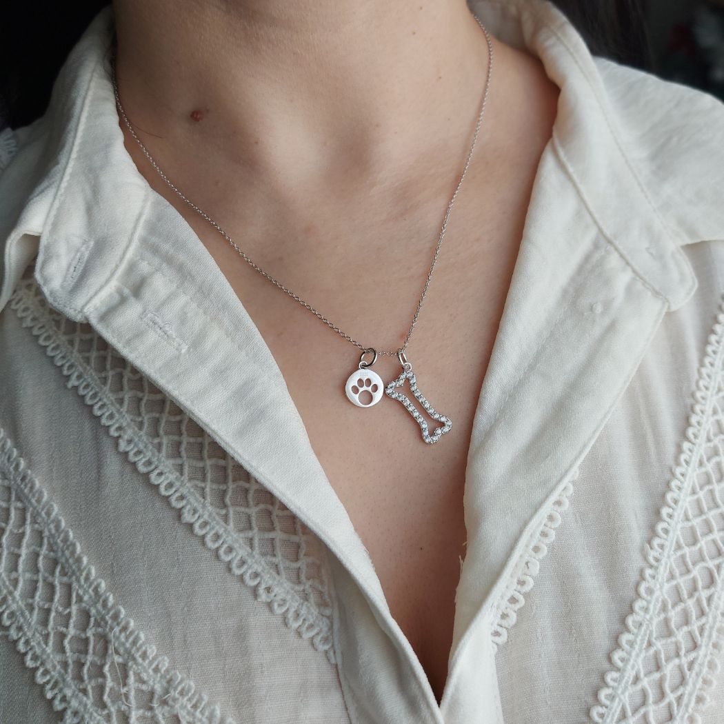 Collar de plata con una huellita dentro de un cÃrculo y un hueso con zirconitas blancas sobre el cuello de una mujer con camisa blanca de el corte inglés