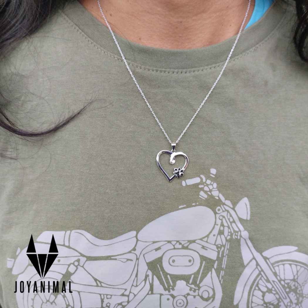 Gargantilla con cadenita fina de plata, sobre cuello de modelo mujer y con colgante con un corazón y una huella negra de gatito o perrito de pandora, sobre camiseta verde, del corte inglés
