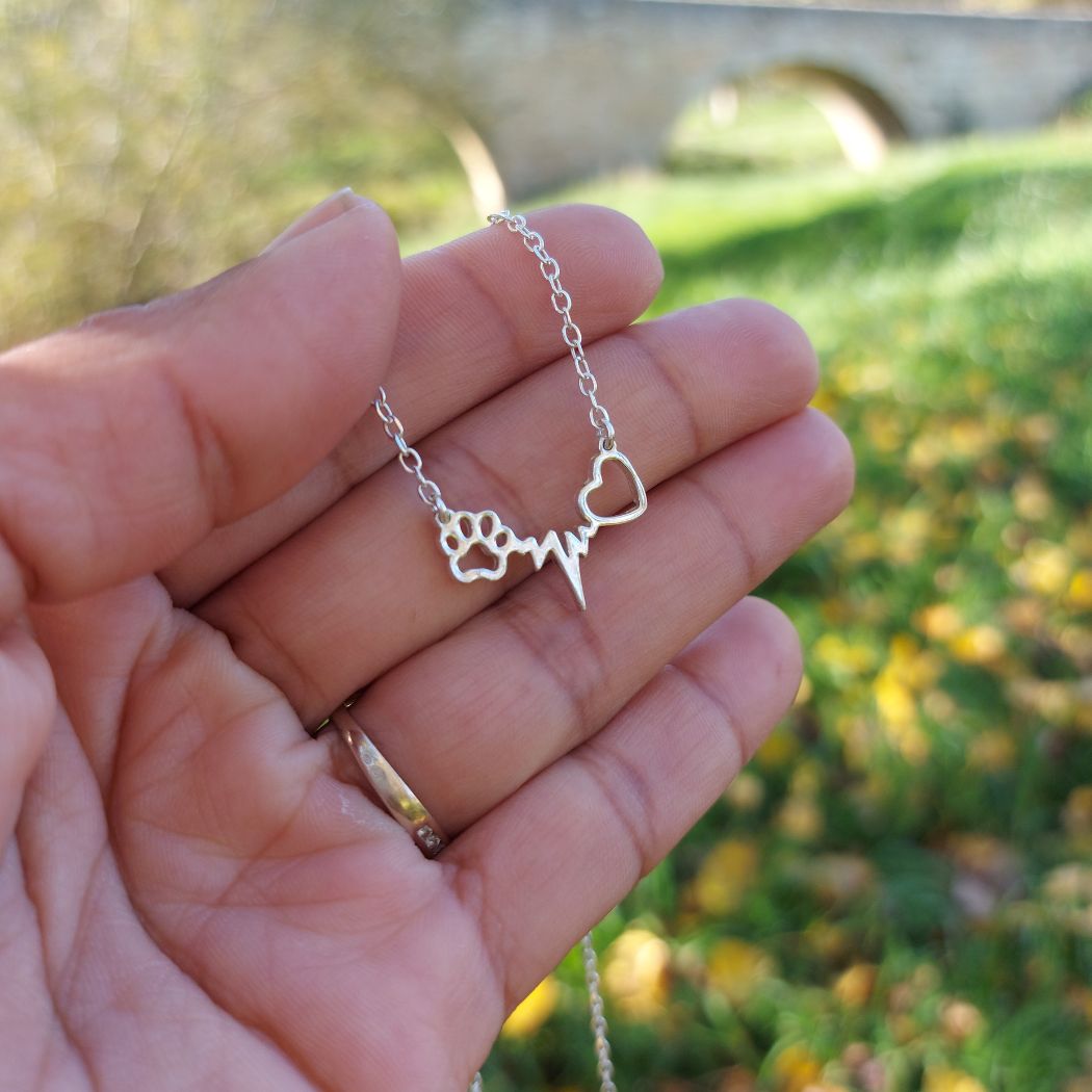 Colgante de plata con forma de huella de perro y gato junto a un corazón y unos latidos, sobre dedos de modelo de san saru