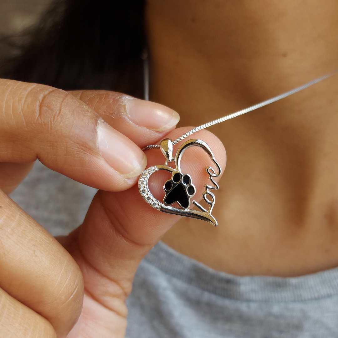 Collar para recordar a tu mascota en el cuello de una mujer y sujetando el colgante en la mano, es un corazón con una huella negra dentro, con brillantes blancos en el contorno y la palabra love de pandora