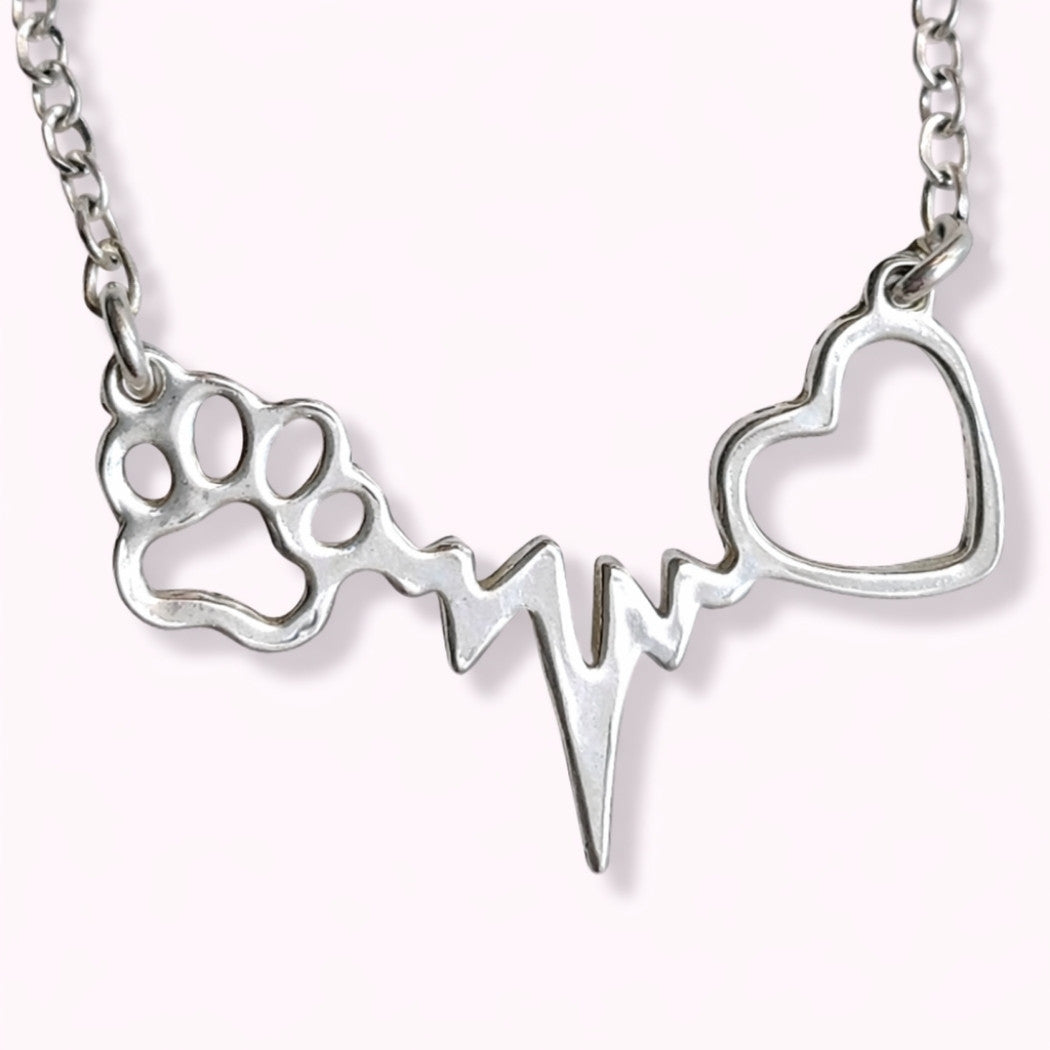 Collar con latidos, una huellita de mascota y un corazón, de plata, sobre fondo rosa pálido de TOUS