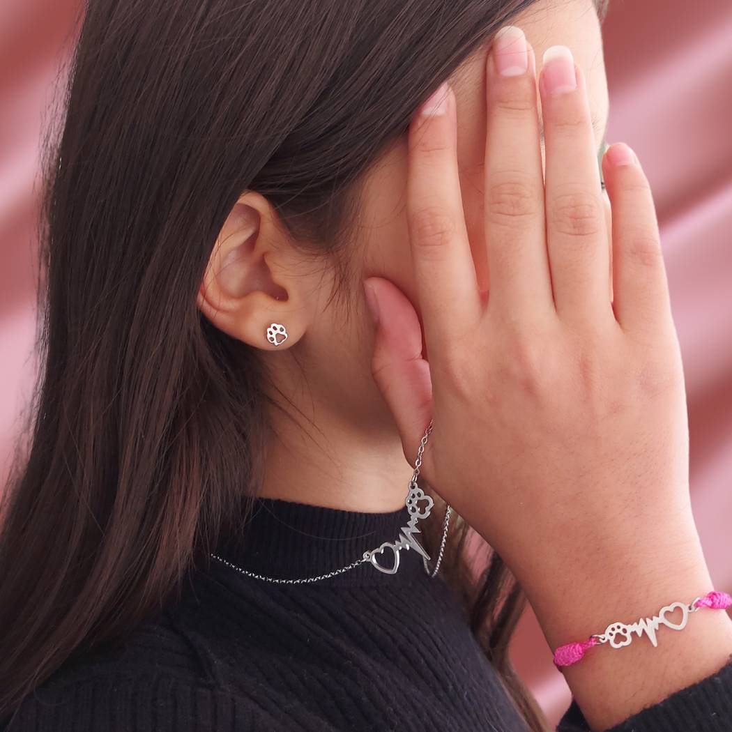 Niña modelando una pulsera rosa de hilo ajustable con colgante de huella y corazon de perro o gato, collar de acero con el mismo diseño que la pulsera y unos pequeños pendientes de plata con huella canina o felina pequeña de TOUS