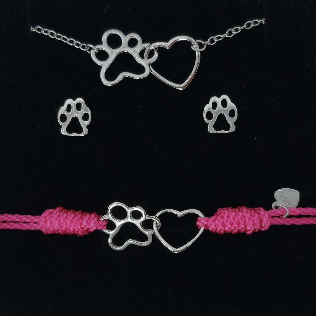 Pulsera de hilo rosa para niña con charm de huella de perro o gato y un corazón de plata, con un collar de plata con colgante a juego de la pulsera y pendientes de plata pequeños con huellita para regalo del corte inglés, sobre fondo negro