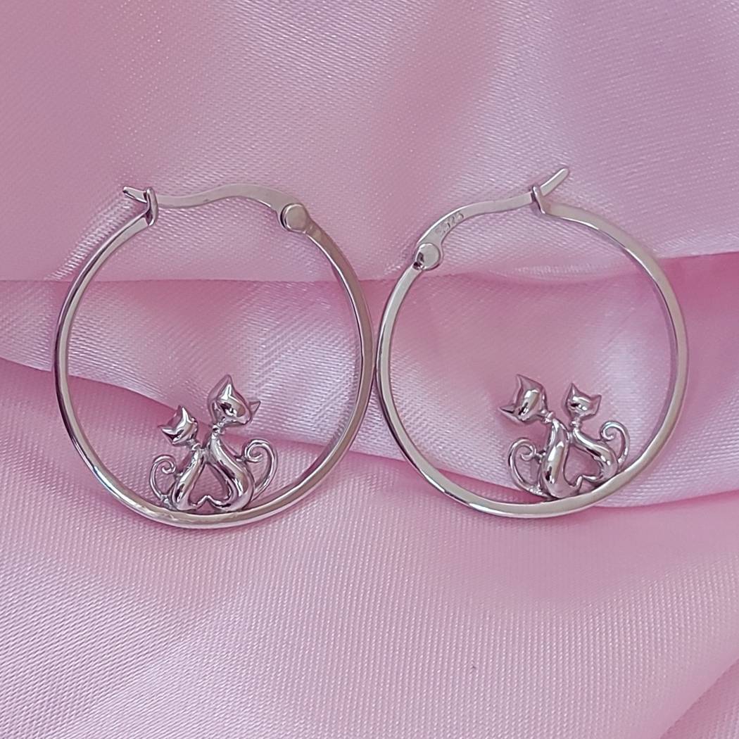 Pendientes de plata con forma de arete con 2 mininos que hacen un corazón con el cuerpo, sobre tela rosa de el corte inglés.