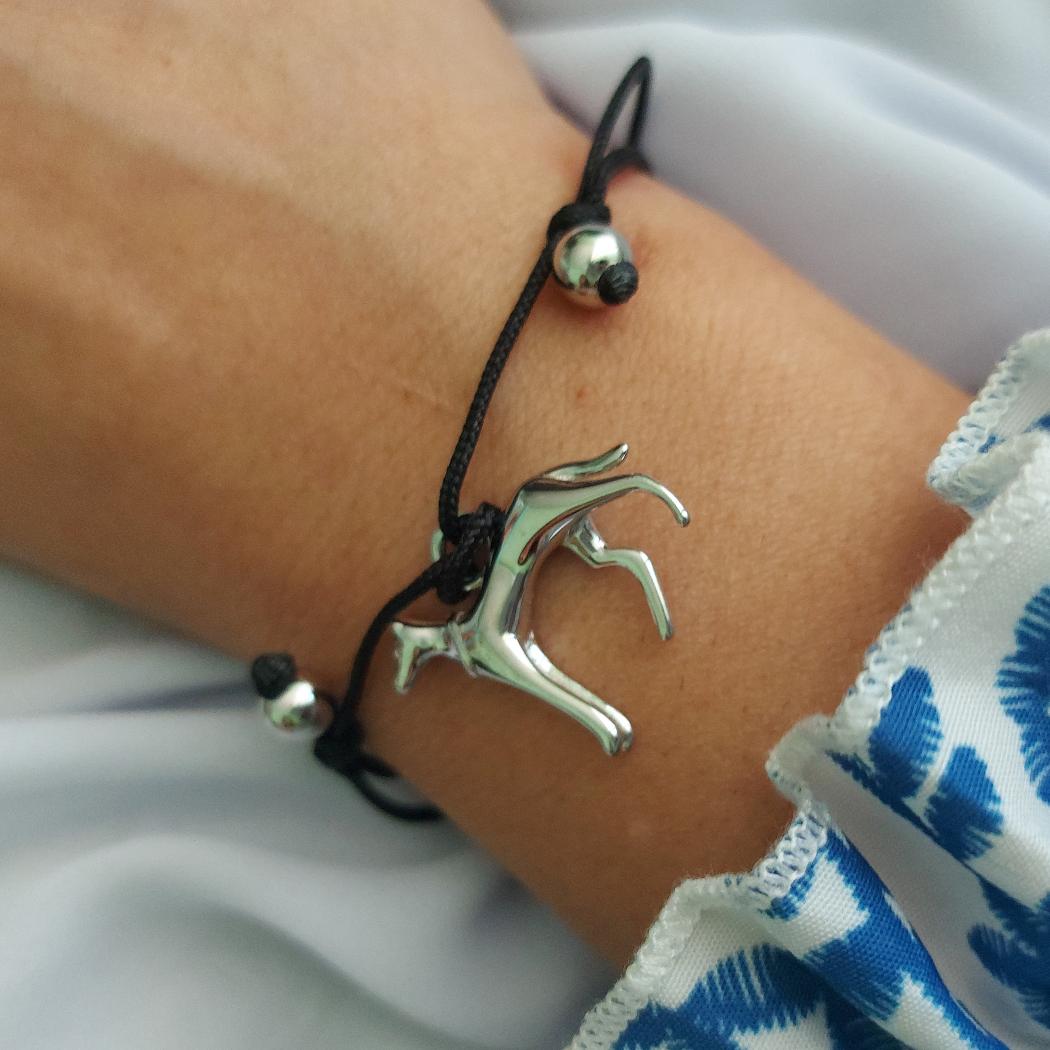 Pulsera con galgo de plata e hilos negros ajustables, en la muñeca de una niña con camisa de flores azules de TOUS