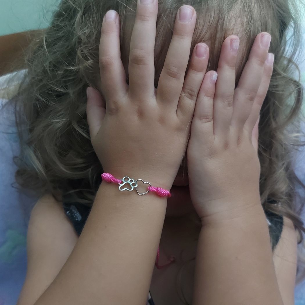 Niña tapándose la cara con las manos, en una muñeca lleva una pulsera ajustable de nudos rosas con un adorno de una huella de mascota y un corazón de TOUS