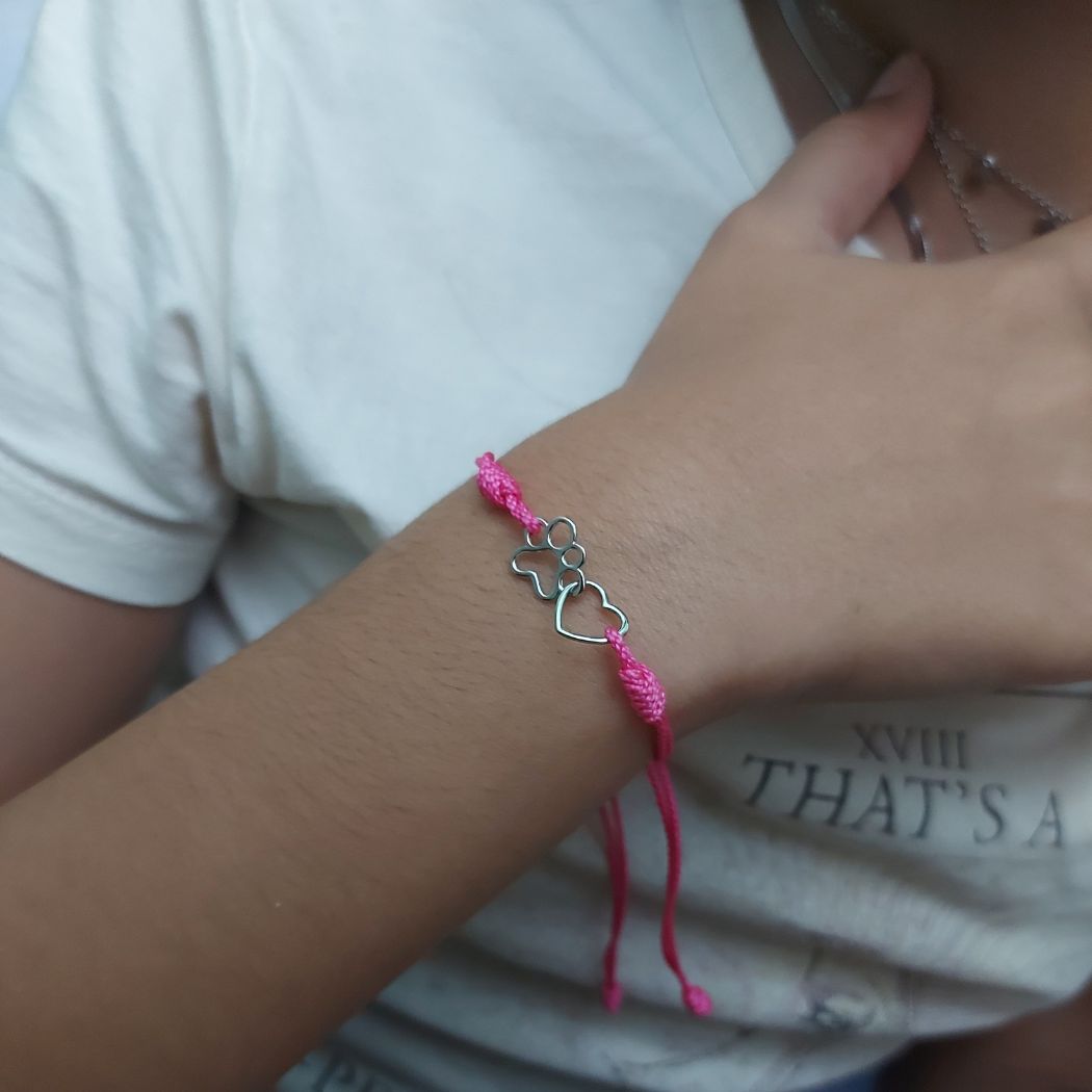 Pulsera con huella de gato y corazón de plata entrelazados con hilo rosa sobre muñeca de mujer con camiseta blanca del corte ingles