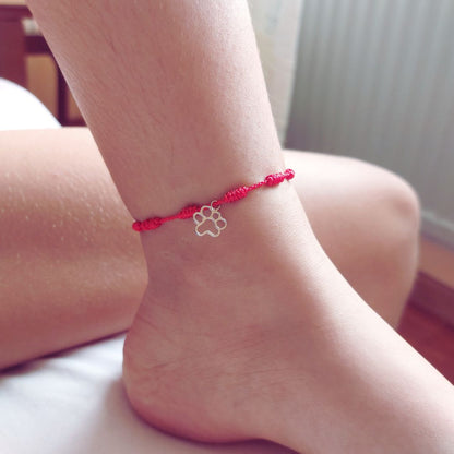 Pulsera roja de la suerte de los 7 nudos puesta en el tobillo como tobillera, lleva un colgante de plata con forma de huella de TOUS