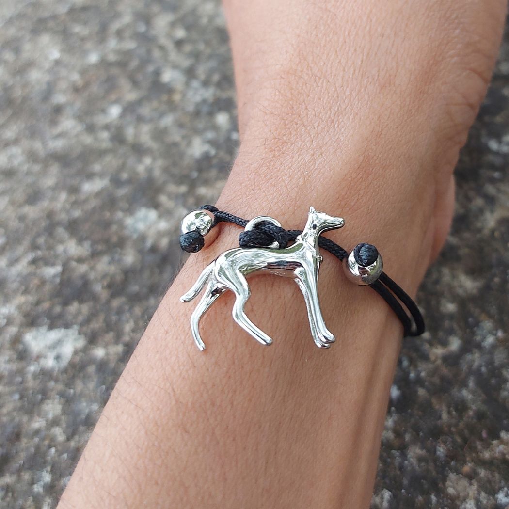 Pulsera de hilo negro con colgante de perro galgo de plata, los nudos ajustables están rodeados con unos adornos de plata en forma de bolitas. Sobre la muñeca de una modelo en una mesa de granito del Corte Inglés.