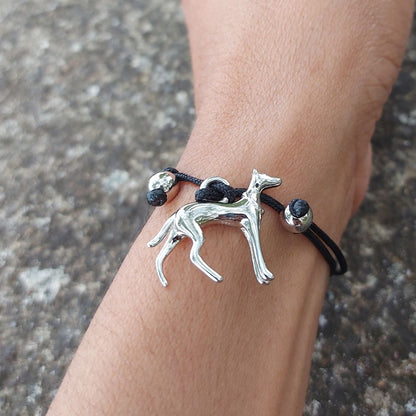 Pulsera de hilo negro con colgante de perro galgo de plata, los nudos ajustables están rodeados con unos adornos de plata en forma de bolitas. Sobre la muñeca de una modelo en una mesa de granito del Corte Inglés.