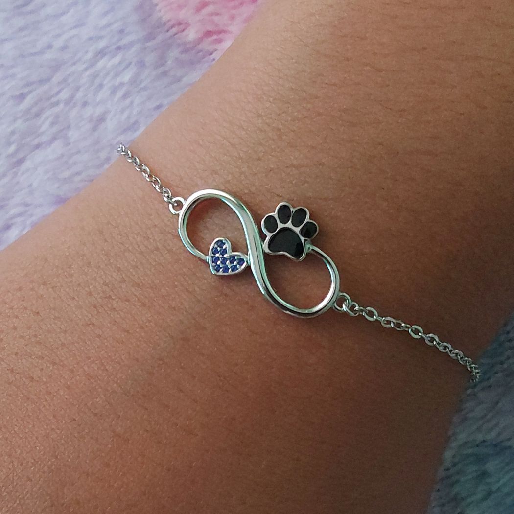 pulsera de plata de un símbolo del infinito con una huellita negra de gato o perro y un corazón con brillantes de zirconia azules, sobre muñeca de mujer, modelo de Pandora