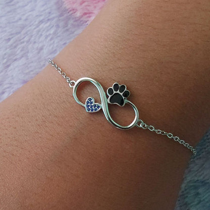 pulsera de plata de un símbolo del infinito con una huella negra de gato o perro y un corazón con brillantes de zirconia azules, sobre muñeca de mujer, modelo de Pandora