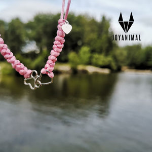 pulsera de nudos de hilo rosa con detalles de huella de mascota y corazón, con un río y árboles de fondo.