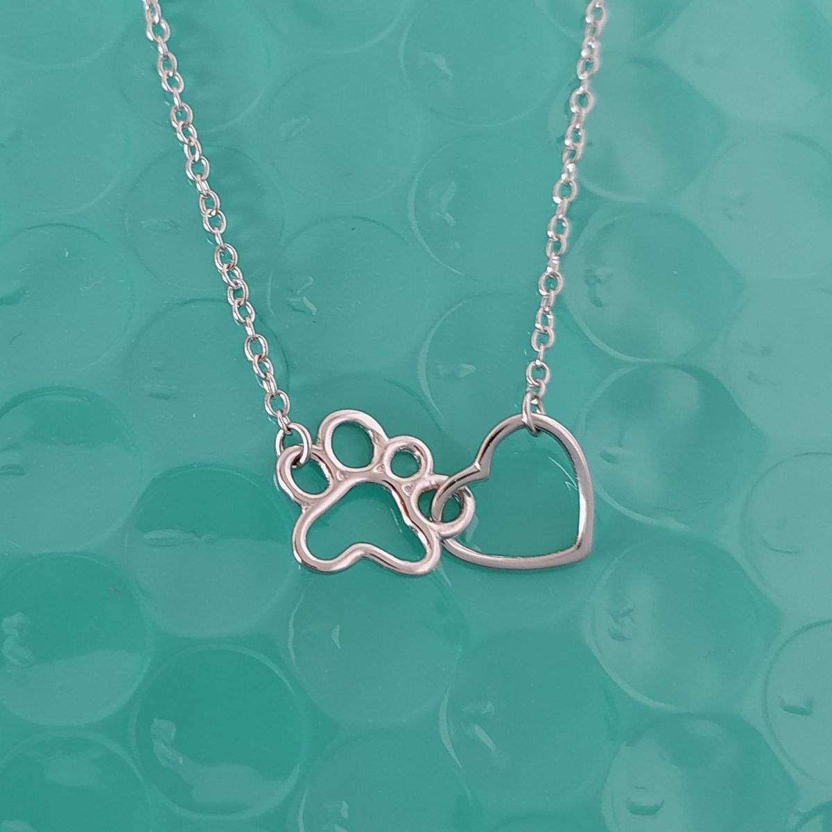 Collar con huellita y corazón en plata de ley, sobre una tela color turquesa del corte ingés