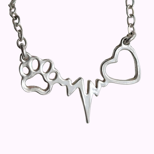 Collar con latidos, una huellita de mascota y un corazón, de plata, sobre fondo rosa pálido de TOUS