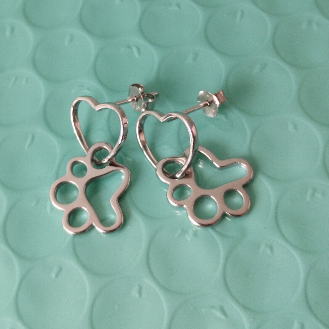 Aretes de plata colgantes con huellas de gato o perro entrelazado con un corazón, sobre fondo con círculos color verde del corte inglés