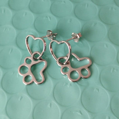 Aretes de plata colgantes con huellas de gato o perro entrelazado con un corazón, sobre fondo con círculos color verde del corte inglés