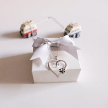 Collar con corazón de plata y huella negra sobre caja de regalo con lacito sobre fondo blanco con 2 juguetes.